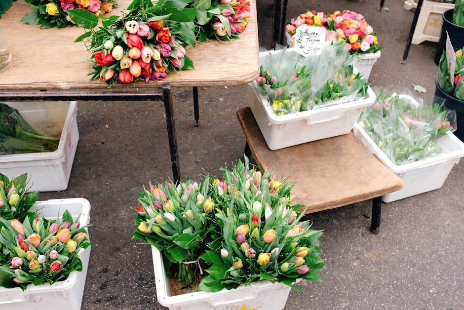 At a flower market, several white plastic crates and wooden tables display a variety of fresh floral arrangements. Prominently, there are two large bouquets of tulips in soft pastel shades of pink, ye