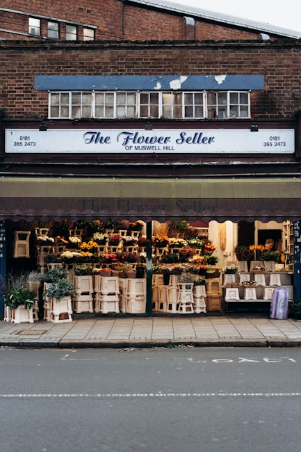 Exterior view of The Flower Seller of Muswell Hill showcasing a display of fresh flowers and floral arrangements arranged on open shelves and white tables beneath a brown awning. The flowers include v