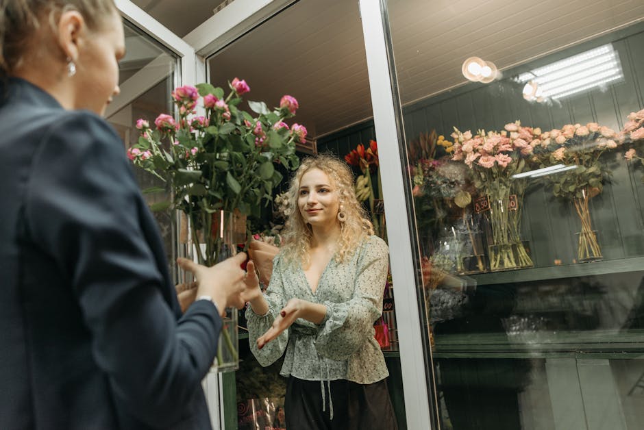 A young woman with curly blonde hair and light skin is inside a florist shop, receiving a bouquet of pink roses with dark green leaves. The bouquet is wrapped in brown paper and held in a glass vase, 