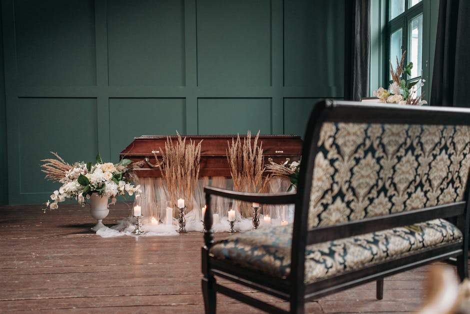 A floral arrangement featuring white orchids, roses, and greenery in a white ceramic vase, placed on a dark wooden table beside a candelabrum with lit candles, complemented by dried pampas grass accen