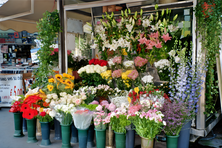 A vibrant display of fresh flowers arranged in various containers outside a floral shop. The arrangement includes white lilies with large petals and green buds, pink lilies with open blooms, and an as
