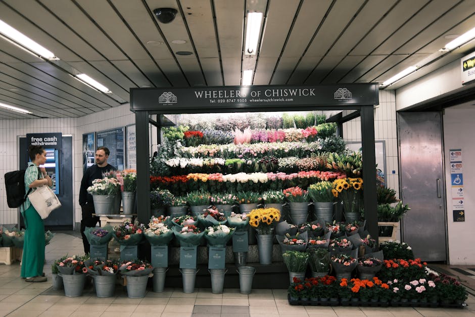 A vibrant flower stand located within a transportation station, featuring a wide variety of fresh flowers arranged in tiered displays. The floral arrangement includes roses, tulips, daisies, and carna